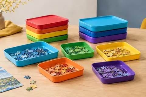 Sept plateaux de tri colorés empilables avec couvercles et pièces de puzzle sur une table en bois clair.